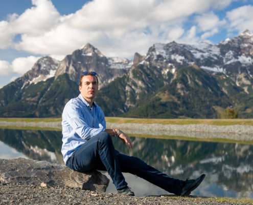 Callboy Linz in blauem Hemd und Jeans sitzt auf einem Felsen an einem ruhigen See, hinter ihm schneebedeckte Gipfel unter einem teilweise bewölkten Himmel.