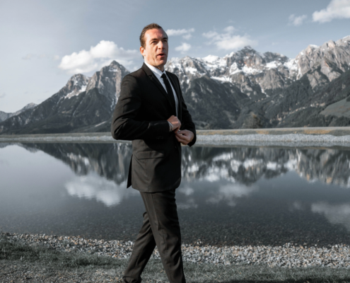 Ein Mann in einem schwarzen Anzug und braunen Schuhen, wie ein Callboy Linz, schlendert auf einem Kiesweg an einem ruhigen See mit schneebedeckten Bergen im Hintergrund.