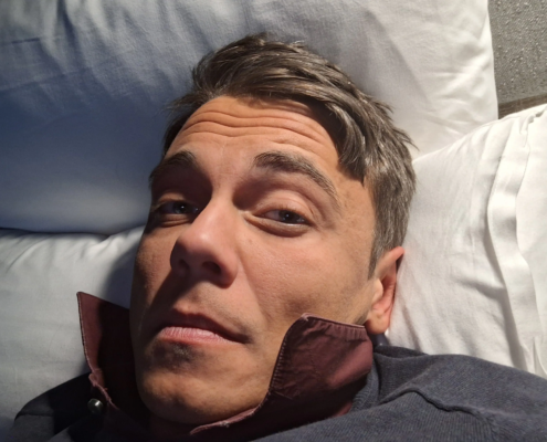 Callboy Tim, kurzes braunes Haar, lehnt in einem dunklen Hemd mit hochgeschlagenem Kragen auf weißen Kissen und blickt neutral in die Kamera.