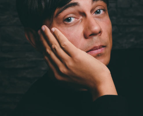 Eine Person mit kurzen dunklen Haaren und hellen Augen, in einem schwarzen Oberteil, stützt ihr Gesicht auf ihre Hand und blickt nachdenklich in die Kamera.