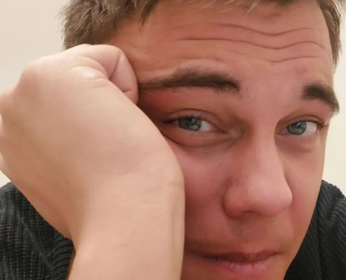 Callboy Tim, kurzes braunes Haar, stützt seinen Kopf auf die Hand und schaut in einem dunklen Pullover mit neutralem oder gelangweiltem Blick in die Kamera.