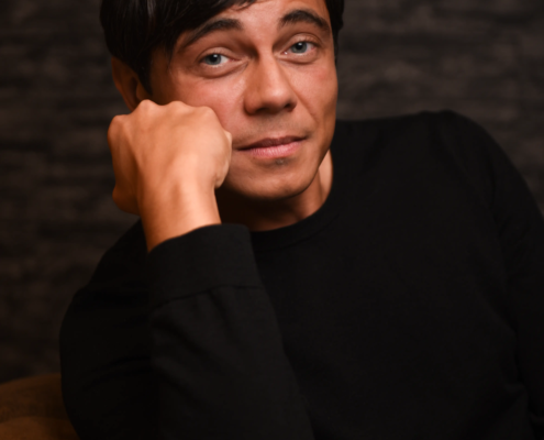 Callboy Tim, kurzes dunkles Haar und helle Augen, in einem schwarzen Hemd, stützt sein Kinn auf die Hand und blickt durch einen dunklen Hintergrund in die Kamera.