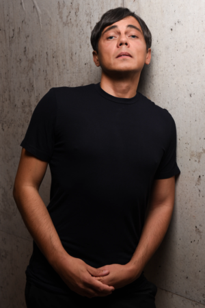 Callboy Tim, kurzes dunkles Haar und schwarzes T-Shirt, steht an einer Betonwand und blickt selbstbewusst mit gefalteten Händen in die Kamera.