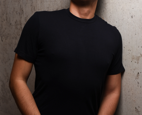 Callboy Tim, kurzes dunkles Haar und schwarzes T-Shirt, steht an einer Betonwand und blickt selbstbewusst mit gefalteten Händen in die Kamera.