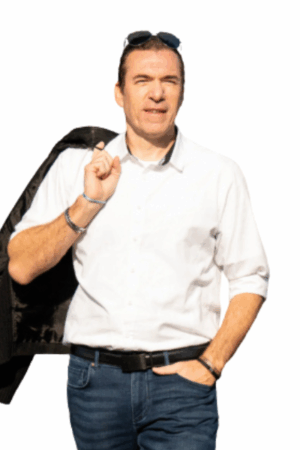 Callboy Chris, in einem weißen Button-up und Jeans mit Sonnenbrille auf dem Kopf, hält eine Jacke über die Schulter vor einem schlichten Hintergrund.