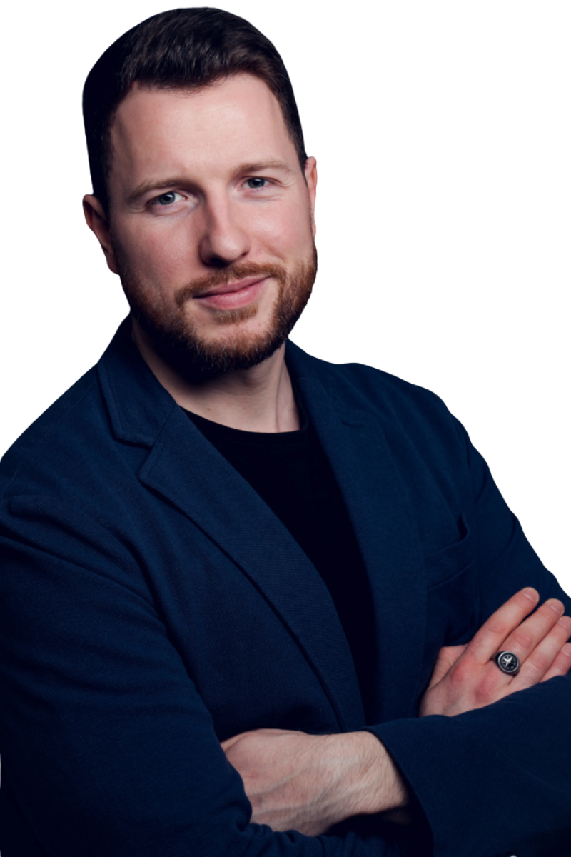 Callboy Georg mit kurzem braunem Haar und gestutztem Bart steht selbstbewusst da, die Arme verschränkt, marineblauer Blazer über schwarzem Hemd, und lächelt.