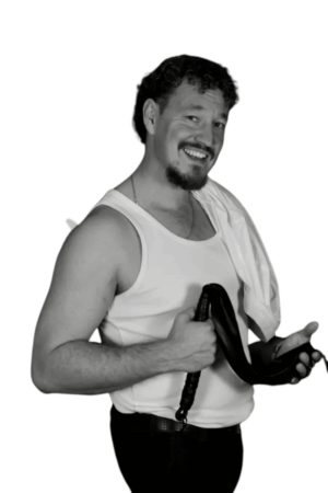 Ein lächelnder Mann mit kurzem lockigem Haar, Spitzbart, Tanktop und Handtuch über der Schulter hält einen Gürtel. Er steht vor einem weißen Hintergrund; schwarz-weißes Bild.