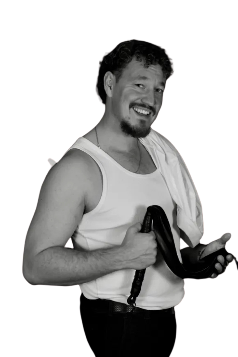 Ein lächelnder Mann mit kurzem lockigem Haar, Spitzbart, Tanktop und Handtuch über der Schulter hält einen Gürtel. Er steht vor einem weißen Hintergrund; schwarz-weißes Bild.