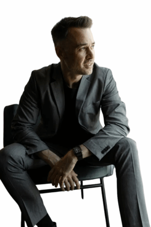 Ein Mann in einem grauen Anzug, der wie Callboy Patrick aussieht, lehnt sich auf einem Stuhl nach vorne, die Hände zwischen den Knien, und blickt nach rechts gegen Weiß.