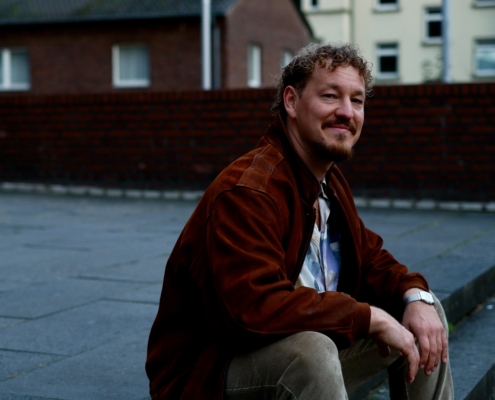 Callboy Jonah, lockig und bärtig, in brauner Jacke und heller Hose, lächelt auf den steinernen Stufen im Freien, dahinter Ziegel und Geländer.