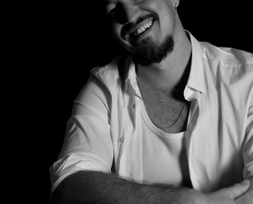 Callboy Jonah, bärtig und mit lockigem Haar, lächelt unter dramatischem Licht in weißem Hemd, Tanktop, Kettenhalsband und Uhr auf Schwarz.