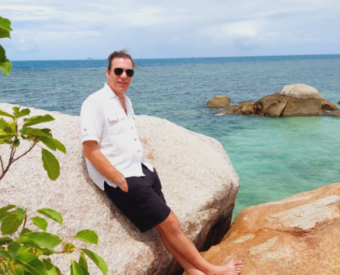 Ein lässiger Mann mit Sonnenbrille, weißem Hemd und schwarzen Shorts liegt barfuß auf den Felsen am Meer, in der Nähe gibt es lebhaftes Wasser und grünes Laub.