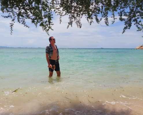 Ein Mann mit Sonnenbrille steht im seichten türkisfarbenen Wasser an einem Sandstrand und entspannt sich unter Zweigen, während der Himmel hell und das Meer ruhig ist.