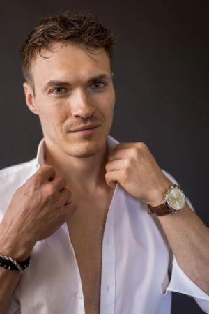 Ein Mann mit kurzen braunen Haaren und hellen Augen fixiert den Kragen seines offenen weißen Hemdes, er trägt eine Uhr und ein schwarzes Armband, der Hintergrund ist dunkel.