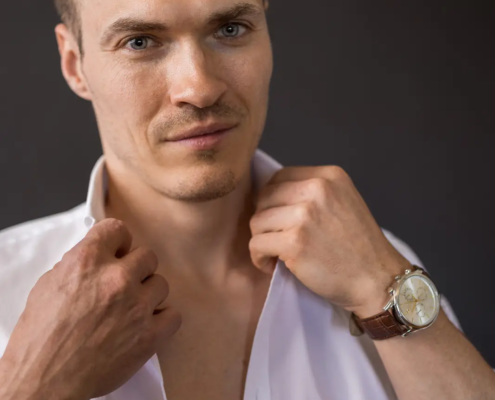 Ein Mann mit kurzen braunen Haaren und hellen Augen fixiert den Kragen seines offenen weißen Hemdes, er trägt eine Uhr und ein schwarzes Armband, der Hintergrund ist dunkel.