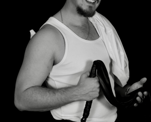Ein lächelnder Mann mit lockigem Haar in einem weißen Tank-Top und einer dunklen Hose hält eine aufgerollte Lederpeitsche, sein weißes Hemd ist über die Schulter gehängt.