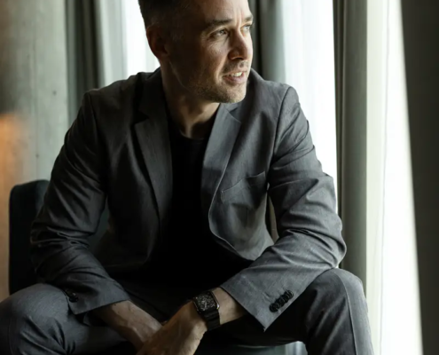 Ein Mann in einem grauen Anzug sitzt auf einem Stuhl am Fenster und blickt nachdenklich hinaus, während weiches Licht durch die Vorhänge sein Gesicht beleuchtet.