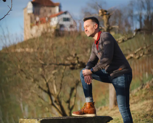 Ein Mann in Jeans und Stiefeln steht mit einem Fuß auf einer Steinbank und lächelt in die Ferne. Das Schloss und die blattlosen Bäume verschwimmen hinter ihm.