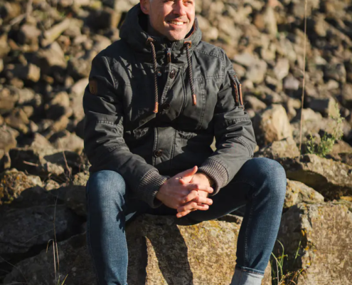 Ein Mann in dunkler Jacke und Stiefeln sitzt lächelnd auf einem sonnenbeschienenen Felsen, umgeben von verstreuten Felsen und einer üppigen grünen Landschaft.