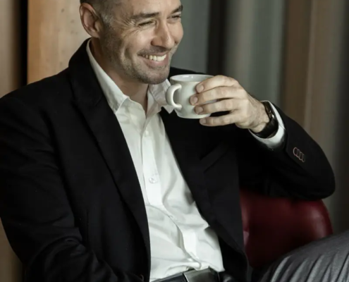 Ein Mann in Anzugjacke und Hemd sitzt entspannt auf einem roten Stuhl, lächelt freundlich und hält eine weiße Kaffeetasse an seinen Mund.