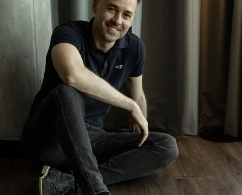 Ein lächelnder Mann mit kurzen dunklen Haaren und Bart sitzt im Schneidersitz auf einem Holzboden, schwarzes Polo und Jeans, dahinter Vorhang und Betonwand.