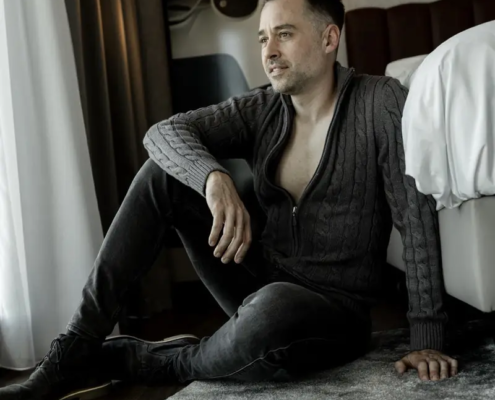 Ein Mann mit kurzen dunklen Haaren in einem grauen Pullover und einer schwarzen Hose sitzt an einem Bett, den Arm auf dem Knie, und schaut nachdenklich zum Fenster.