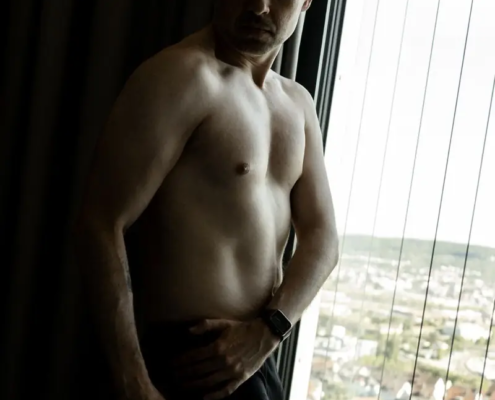 Ein Mann ohne Hemd und in schwarzer Unterwäsche steht an einem Fenster mit durchsichtigen Vorhängen und blickt nach unten. Draußen erscheinen Sonnenlicht und eine Stadtlandschaft.