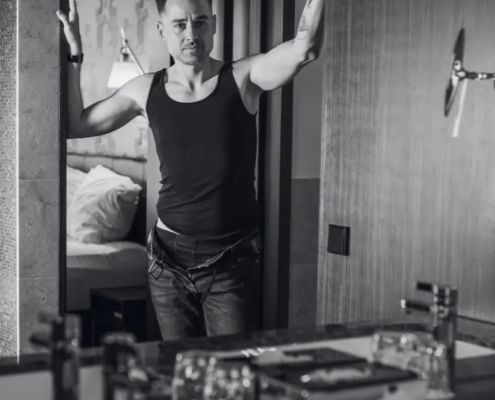 Ein Mann in einem schwarzen Tank und Jeans steht in einer Tür, die Arme weit ausgebreitet, mit ernster Miene. In einem Innenraum in der Nähe eines Waschbeckens. Schwarz-Weiß-Bild.