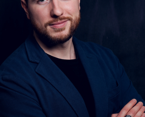 Ein bärtiger Mann mit kurzen braunen Haaren in einem dunkelblauen Blazer und einem schwarzen Hemd steht mit verschränkten Armen da und blickt selbstbewusst in die Kamera.