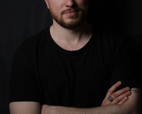 Ein Mann mit kurzen Haaren und Bart steht vor einem dunklen Hintergrund in einem schwarzen T-Shirt, die Arme verschränkt, mit einem leichten Lächeln in die Kamera blickend.
