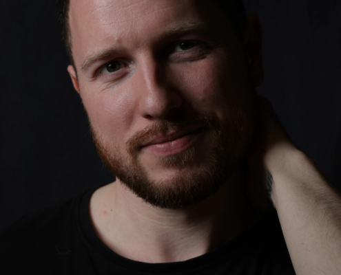 Ein bärtiger Mann mit kurzen braunen Haaren in einem schwarzen Hemd lächelt sanft, die rechte Hand im Nacken, das Gesicht beleuchtet vor einem dunklen Hintergrund.