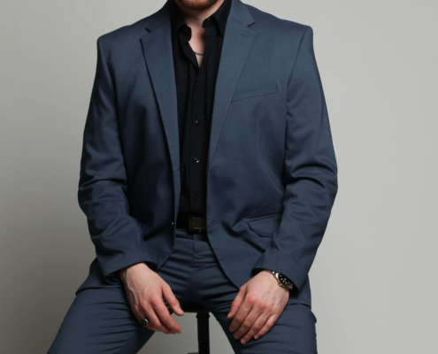 Ein lächelnder Mann mit kurzen Haaren und Bart in einem blauen Anzug und schwarzem Hemd sitzt auf einem schwarzen Hocker vor einem schlichten hellgrauen Hintergrund.