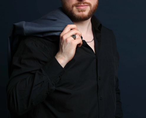 Vor einem dunklen Hintergrund steht ein Mann mit kurzen braunen Haaren und einem Bart, der ein schwarzes Hemd trägt und seine Jacke über eine Schulter hält.