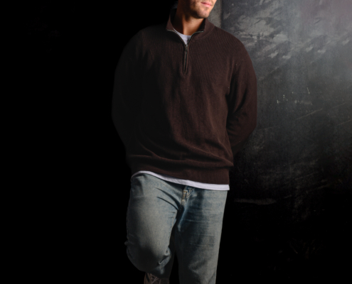 Callboy Tim, bekleidet mit einem dunklen Pullover und Jeans, lehnt mit einem gekreuzten Bein an einer schattigen Wand, das Licht auf seiner rechten Seite.