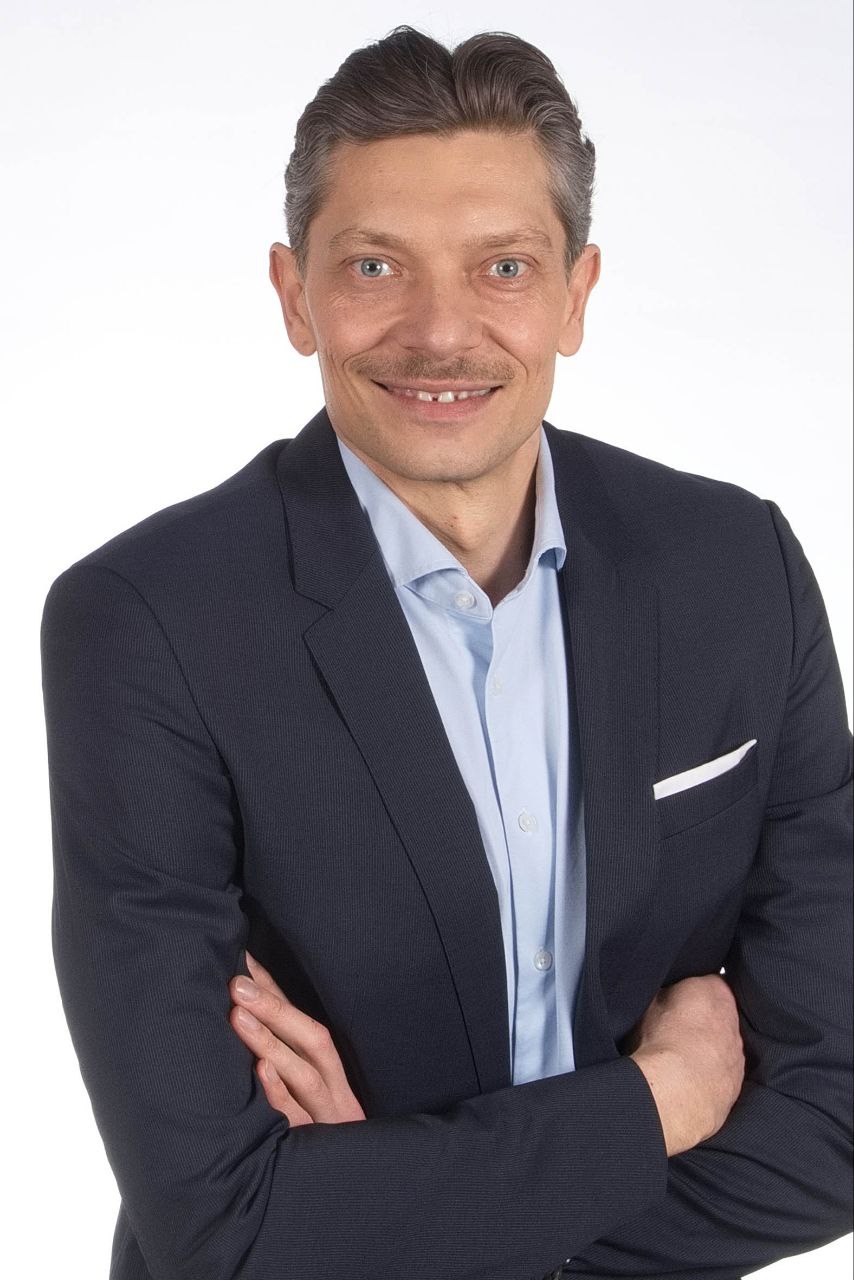 Callboy Jim, im dunklen Anzug und hellblauem Hemd, steht mit verschränkten Armen und selbstbewusstem Lächeln vor einem schlichten weißen Hintergrund in die Kamera.