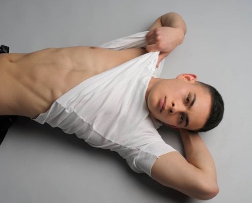 Bruno liegt auf dem Rücken und hebt sein weißes T-Shirt an, um seinen straffen Bauch zu zeigen. Er trägt eine graue Hose und kurze dunkle Haare.