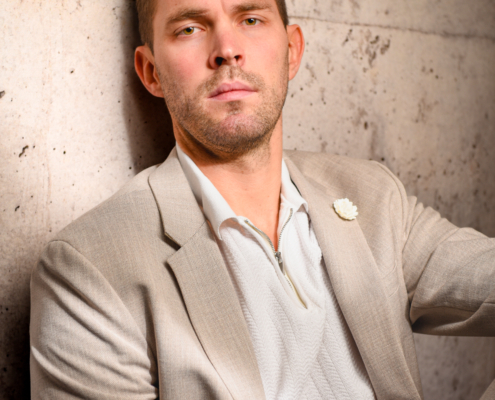 Callboy Tim, kurzhaarig mit gepflegtem Bart, im beigen Anzug und weißem Hemd, sitzt an einer Steinmauer und blickt selbstbewusst nach vorn.