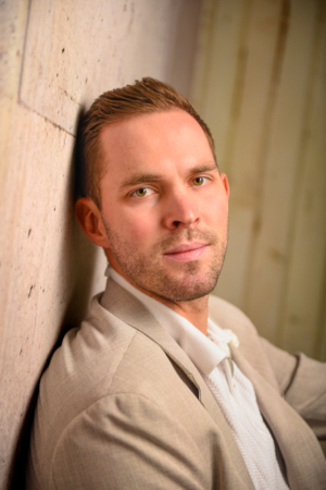 Callboy Tim, mit kurzen braunen Haaren und gepflegtem Bart, in beiger Jacke und weißem Hemd, lehnt an einer Steinmauer und blickt ruhig vor sich hin.