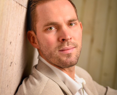 Callboy Tim, mit kurzen braunen Haaren und gepflegtem Bart, in beiger Jacke und weißem Hemd, lehnt an einer Steinmauer und blickt ruhig vor sich hin.