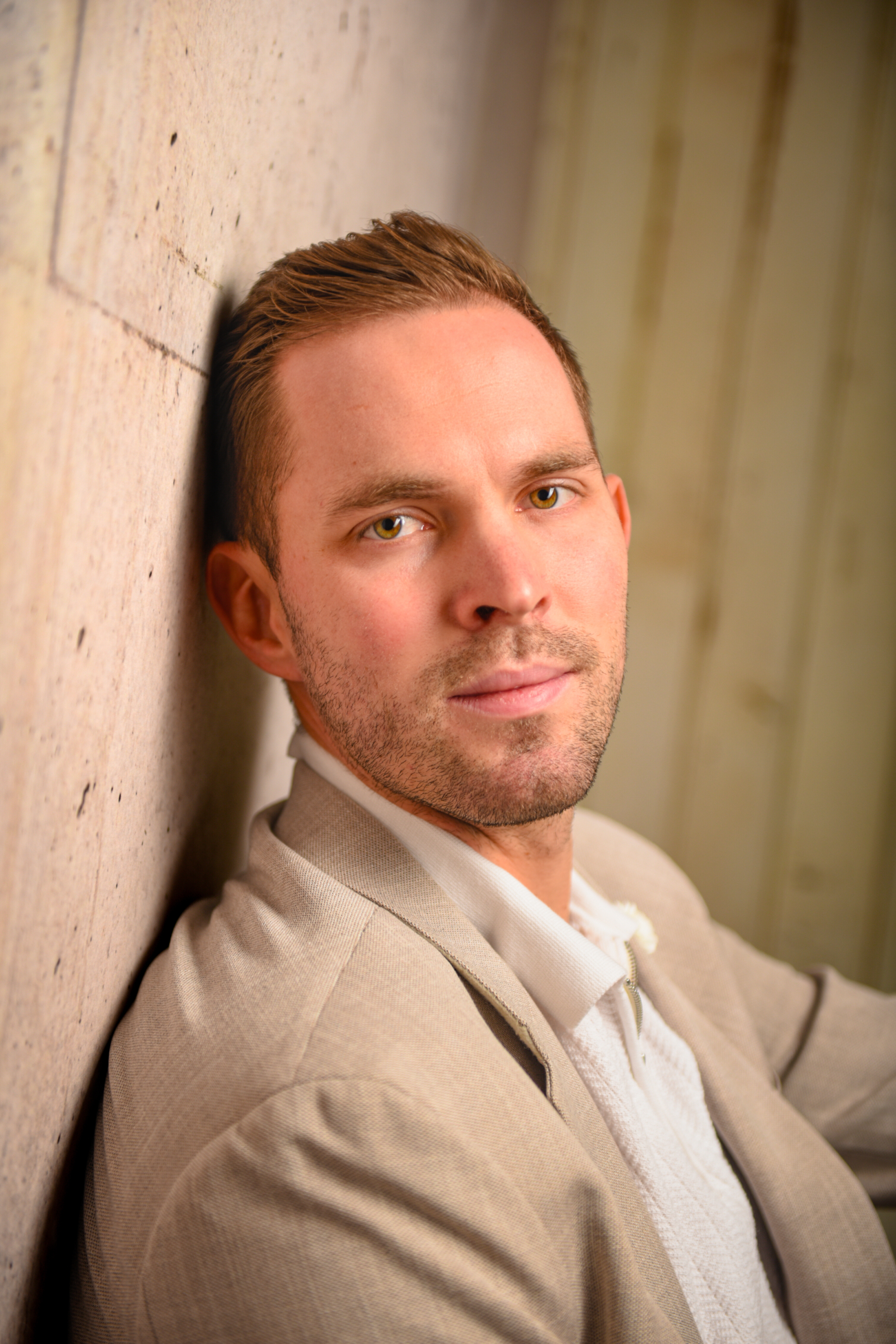 Callboy Tim, mit kurzen braunen Haaren und gepflegtem Bart, in beiger Jacke und weißem Hemd, lehnt an einer Steinmauer und blickt ruhig vor sich hin.