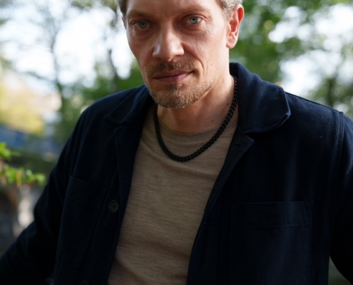 Ein Mann mit kurzen, gewellten braunen Haaren und leichten Bartstoppeln steht draußen in einem beigen Hemd und einer dunklen Jacke, eine Halskette ist sichtbar. Bäume und Sonne im Hintergrund.