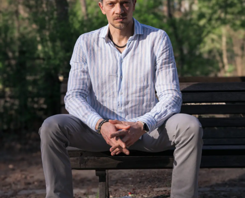 Callboy Jim, mit kurzen Haaren, Schnurrbart und hellem Bart, sitzt auf einer Parkbank in blauen Streifen, während das Sonnenlicht durch die Bäume fällt.