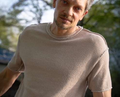 Ein Mann mit kurzen braunen Haaren und einem gestutzten Bart, wie Callboy Jim, sitzt in einem beigen Hemd im Freien. Hinter ihm stehen sonnenbeschienene grüne Bäume.