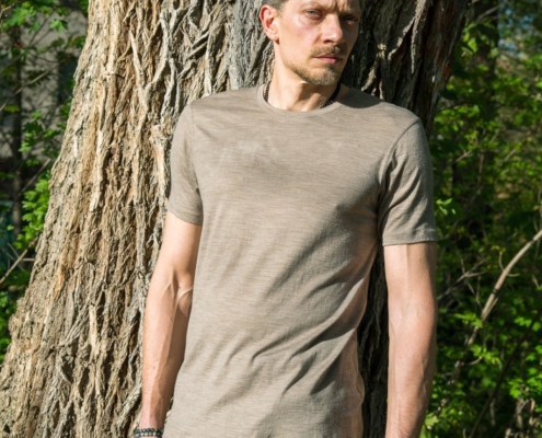 Callboy Jim, ein Mann mit kurzen hellbraunen Haaren, steht draußen an einem Baum in einem beigen T-Shirt und einer grauen Hose, die vom Sonnenlicht beschienen wird.