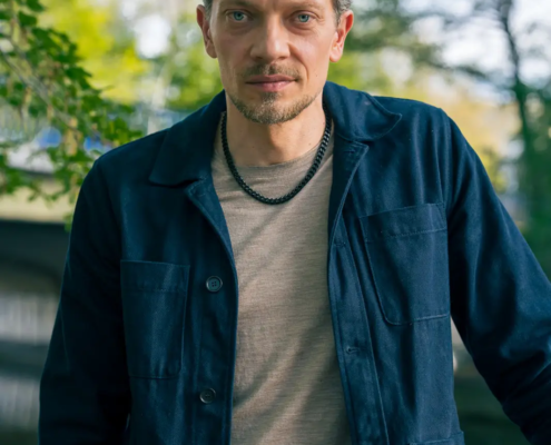 Callboy Jim, mit kurzen, gewellten braunen Haaren und hellen Augen, steht draußen in einer marineblauen Jacke, einem beigen Hemd und schwarzen Perlen inmitten sonnenbeschienener Bäume.