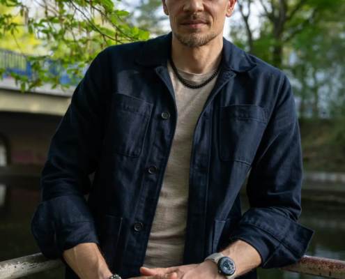 Ein bärtiger Mann mit kurzem, gewelltem Haar steht in einer dunklen Jacke und einem beigen Hemd an einem Geländer im Freien und blickt auf Wasser und Bäume.