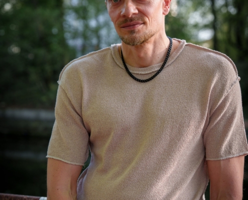 Callboy Jim, hellbraunes Haar und kurzer Bart, in beigefarbenem, kurzärmeligem Hemd und schwarzer Halskette, lehnt draußen im Sonnenlicht an einem Geländer.