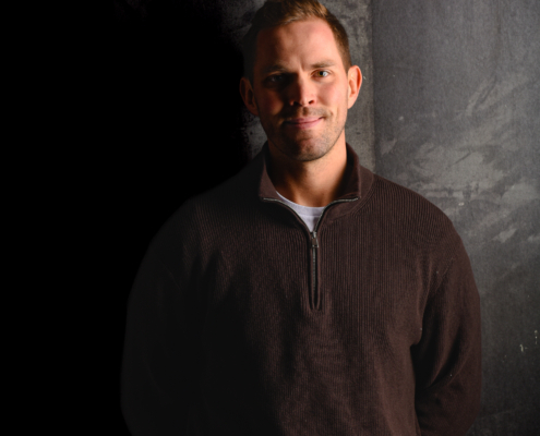 Callboy Tim, mit kurzen hellbraunen Haaren, in einem dunklen Pullover mit Viertelreißverschluss und hellen Jeans, lächelt sanft in dramatischem Licht.