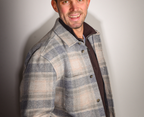 Callboy Tim, kurze braune Haare, trägt eine grau-beige karierte Jacke über einem dunklen Hemd und lächelt in die Kamera vor einem hellen Hintergrund.