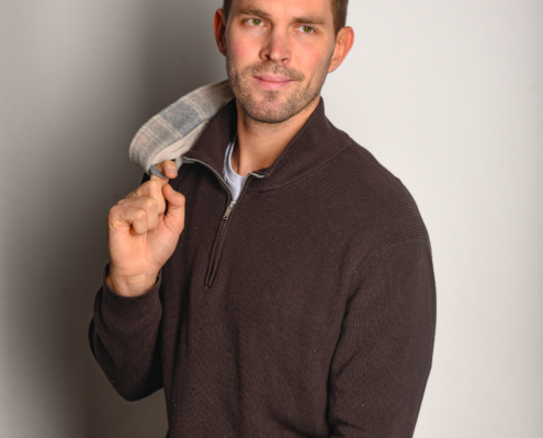 Callboy Tim, kurzes braunes Haar und gestutzter Bart, sitzt auf einem Hocker in Pullover, Jeans, karierter Jacke über der Schulter, leichtes Lächeln.